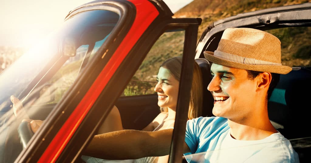 A man and woman in a red car smiling under the sunshine.