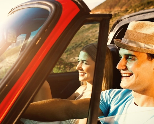 A man and woman in a red car smiling under the sunshine.