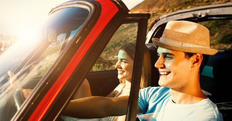 A man and woman in a red car smiling under the sunshine.