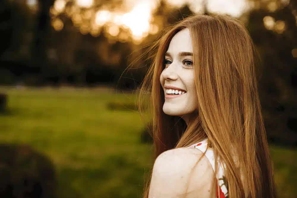 A woman with long red hair smiling in a park, showcasing the successful results of a hair restoration treatment plan.