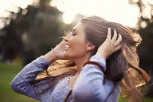A woman with her hands on her hair is smiling in a park.